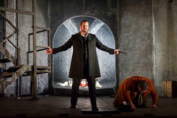 Don Giovanni is standing centre stage, arms wide, with a gun in his left hand. Leporello is on the floor bent over on stage left.