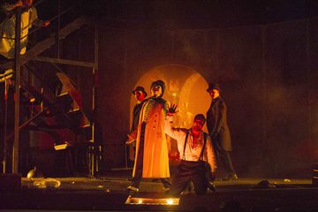 Don Giovanni is on his knees and Il Commendatore is standing above him. Il Commendatore is holding Don Giovanni's hand above a fire pit in the middle of the stage