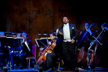 A man stands in full black tie singing in front of the cellos and double basses of the orchestra