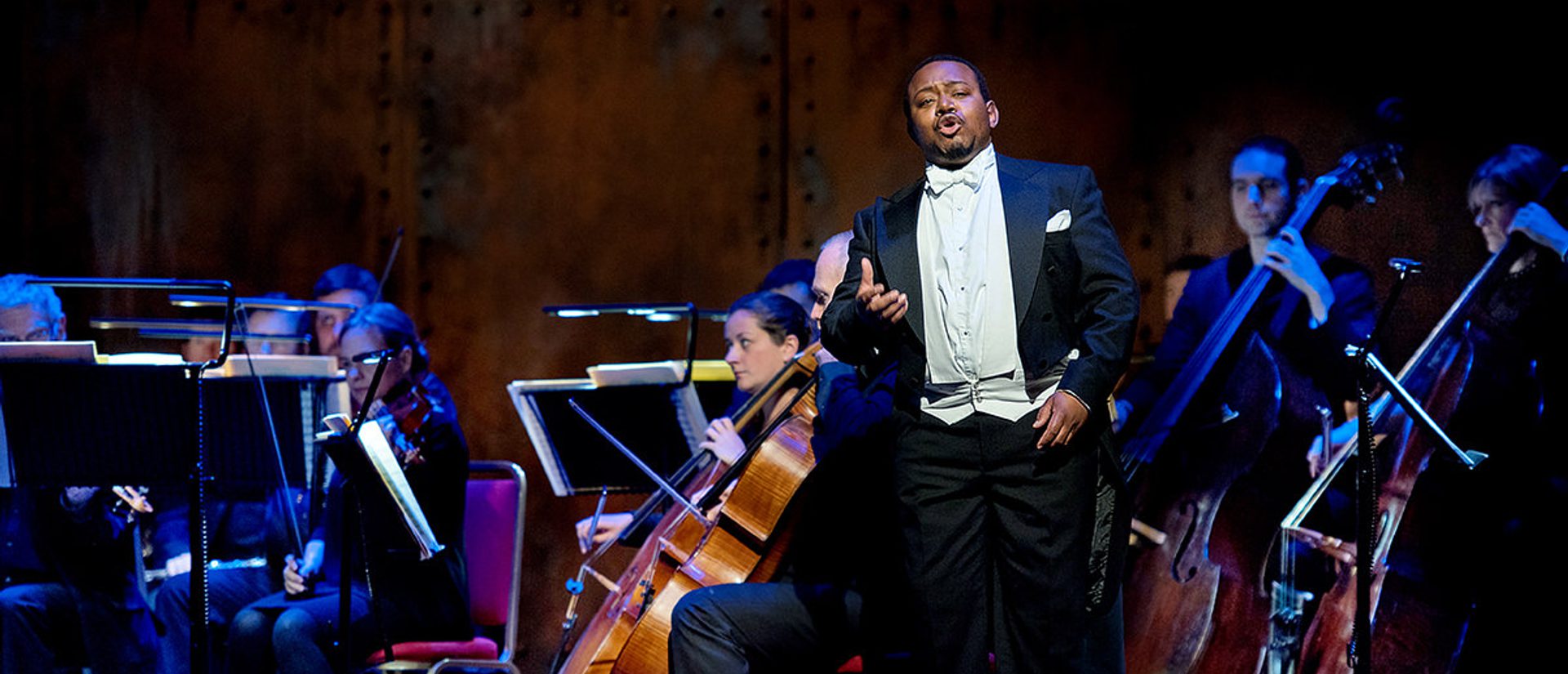 A man stands in full black tie singing in front of the cellos and double basses of the orchestra