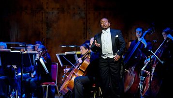 A man stands in full black tie singing in front of the cellos and double basses of the orchestra