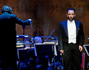 A man stands in full black tie, singing. There is a conductor and an orchestra in the background