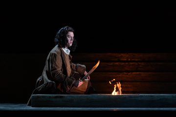Achilla kneels by fire, holding his sword against the flame