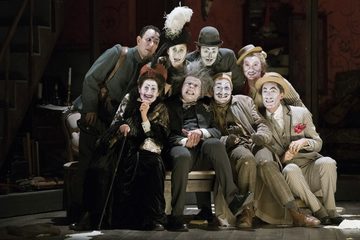 A group is huddle round Simone. Their faces are painted in a grotesque style and they are all looking out into the distance