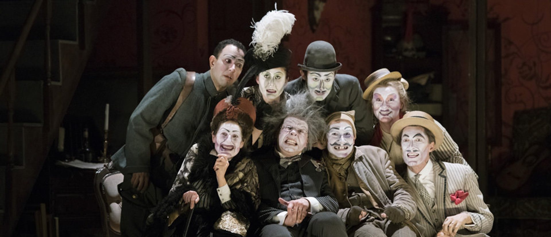A group is huddle round Simone. Their faces are painted in a grotesque style and they are all looking out into the distance