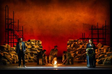 Male and Female chorus stand on either edge of the stage, looking at each other with concern. In the center of the stage, 3 performers dressed in military suits sit around a fire.