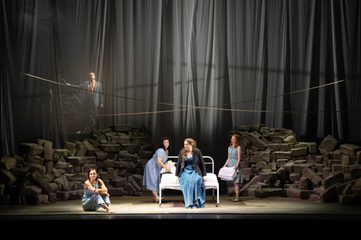 Female Chorus, Bianca and Lucia stand around a bed in the center of the stage and look over to Lucretia sitting on the floor. Male Chorus stands menacingly at the back of the stage behind a sheer curtain.