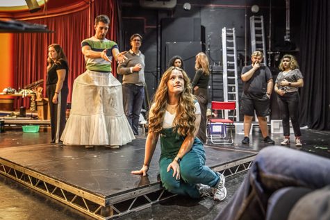 A woman in overalls sits on the front of a stage. Behind her, a man wearing a skirt gestures towards her.