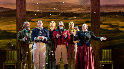 A group of singers stand on a dimly lit stage, looking upwards, as petals fall from into the frame.