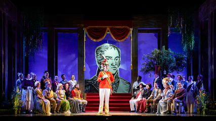 The cast of Il viaggio a Reims sit in two sets of rows, watching a male wearing a red, period uniform, sing. Behind them is a grand backdrop featuring an image of King Charles X.