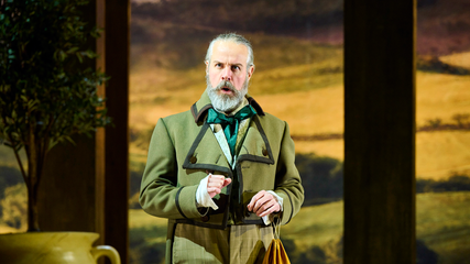 A male singer, wearing a green overcoat and silk green neck tie, stands against a dimly lit forest backdrop. He is holding a bag, and has his arms in front of him.