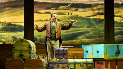 A male singer, wearing a brown period suit and glasses stands in front of multiple old luggage pieces. He is gesturing towards the audience, singing.