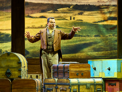 A male singer, wearing a brown period suit and glasses stands in front of multiple old luggage pieces. He is gesturing towards the audience, singing.