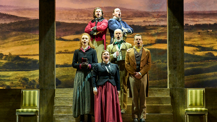 A group of singers stand on steps, against a woodland backdrop. They are all singing.