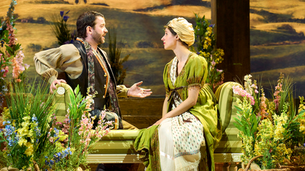 A male and female sit on a green sofa, surrounded by flowers. The male is gesturing to the female.