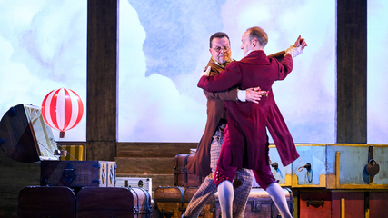 Two male singers are dancing together, in front of a backdrop that shows a blue sky. To their right is a small hot air balloon, appearing out of a piece of luggage.