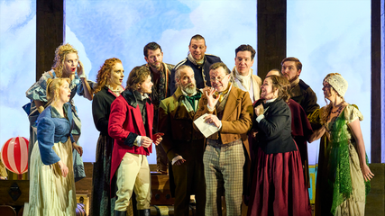 The cast of Il Viaggio a reims stand against a sky background, huddled around one male singer who is holding a piece of paper, and is singing.