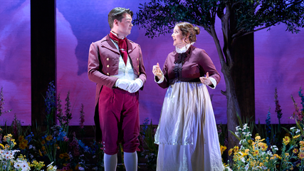A male and female singer stand against a purple sky backdrop, surrounded by flowers. They are wearing burgundy hotel uniforms, and are singing.