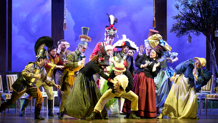 The cast of Il Viaggio a Reims are stood centre stage, in front of a purple sky backdrop. They are wearing extravagant period clothing and are all leaning to the right.