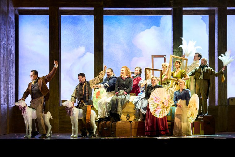 Men and women in period dress form the shape of a carriage on stage in front of a cloud backdrop.