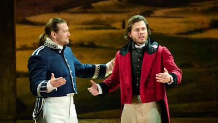 Two male singers are standing against a field backdrop. One is wearing a blue suit jacket, and is holding the arm of the other, who is wearing a red suit jacket and is looking out to the audience.