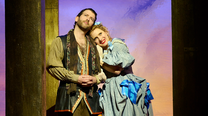 A male and female wearing period clothing stand against a sunset background. The female has her head rested on his shoulder, smiling.