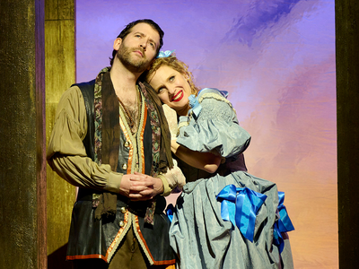 A male and female wearing period clothing stand against a sunset background. The female has her head rested on his shoulder, smiling.